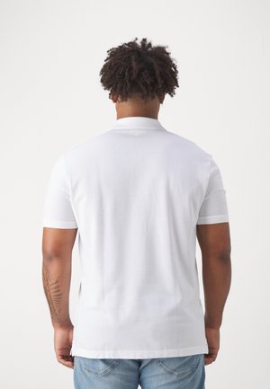 Mann mit lockigem Haar, der ein schlichtes weißes kurzärmeliges Hemd und hellblaue Jeans trägt, von hinten auf weißem Hintergrund gesehen.