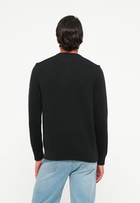 Hombre de espaldas, vistiendo un suéter negro de manga larga y jeans azul claro contra un fondo blanco liso.