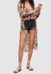 Květinový dlouhý přehoz z průsvitné látky s růžovými, červenými a zelenými vzory, doplněný černými denimovými šortkami a béžovými sandály na řemínky.