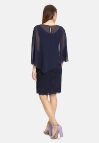 Femme portant une robe en dentelle bleu marine longueur genou avec des manches cape transparentes bleu marine, associée à des sandales à talons hauts lavande avec bride à la cheville, debout de dos.