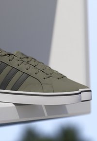 Par de zapatillas verde oliva con rayas negras y suelas blancas descansando sobre un borde blanco contra un fondo gris y blanco.