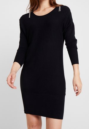 Kobieta ubrana w czarną, dzianinową sukienkę z długim rękawem, z prążkowanym dołem i mankietami, ozdobioną drobnymi kryształkami na ramionach.