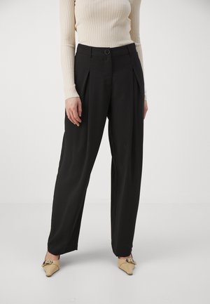 Femme portant un pantalon large noir, un haut beige à manches longues côtelé et des chaussures beiges pointues avec un détail de boucle, debout sur fond blanc.