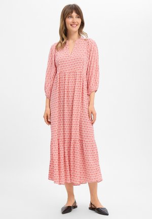 Vrouw glimlachend, dragend een midi-jurk met lange mouwen in roze-wit patroon en zwarte platte puntige slingback schoenen op een witte achtergrond.