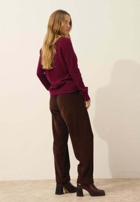 Burgundin neulepaita, joka on yhdistetty ruskeisiin samettihousuihin, joissa on rento istuvuus. Mallilla on tummat korkokengät. Yksinkertainen tausta.