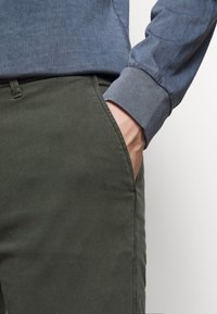 Olijfgroene broek van een gestructureerde stof, rechte snit, en zijzakken; vergezeld door een blauwe, lange mouw, geknoopt shirt.