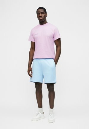 Mężczyzna stojący w jasnofioletowej koszulce, jasnoniebieskich szortach, białych skarpetkach i białych trampkach na jednolitym jasnym tle.