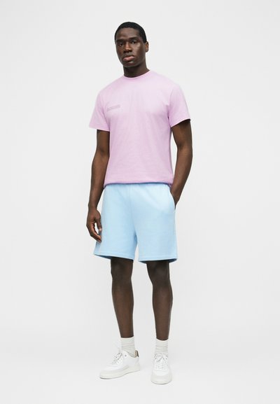 Homme debout portant un t-shirt violet clair, un short bleu clair, des chaussettes blanches et des baskets blanches, sur un fond clair uni.