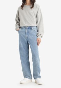 Sudadera gris de manga larga con el logo 'LEVIS', combinada con unos jeans rectos de color azul claro y zapatillas blancas, de pie frente a un fondo liso.