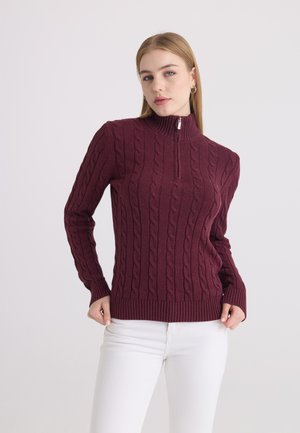 Jeune femme portant un pull torsadé bordeaux avec un col à zip partiel et un pantalon blanc, debout devant un fond uni.