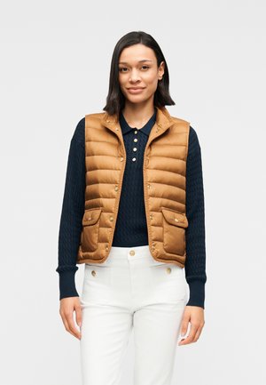 Brun polstret vest med høj krave og trykknapper, der har to forlommer foran. Båret over en navyblå strikket sweater og hvide bukser.