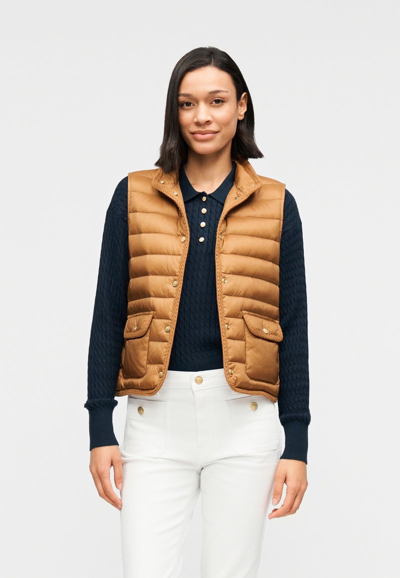 Καφέ μονοκέφαλο γιλέκο με ψηλό γιακά και κουμπιά με κλικ, με δύο μπροστινές τσέπες. Φορεμένο πάνω από ένα σκούρο μπλε πλεκτό πουλόβερ και λευκό παντελόνι.