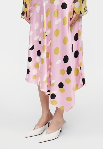 Ružové šaty s viacfarebnými (zlatými, bielymi, čiernymi) bodkami, s prúdiacim asymetrickým lemom, doplnené bielymi lodičkami s výraznými špičkami a čipkovanými detailmi.
