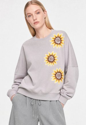 Kvinde iført en lysegrå sweatshirt med tre store solsikkemotiver foran og matchende grå bukser med snore.