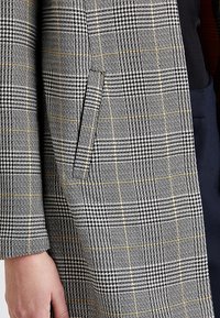 Abrigo de cuadros gris con un tejido texturizado, con un sutil acento amarillo y un bolsillo lateral en la manga izquierda.