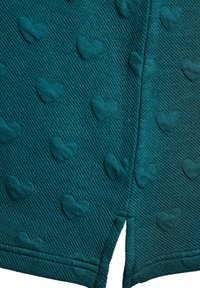 Tyrkysově modré kraťasy z tkaniny s texturovaným vzorem malých vystouplých srdíček a postranním rozparkem. Materiál má žebrovaný vzhled pro jedinečný efekt.