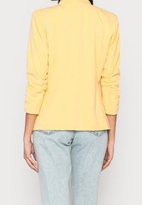 Blazer jaune cintré à manches longues, détails froncés aux manches, porté avec un jean bleu clair, texture lisse, design minimaliste.