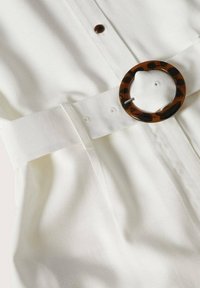 Ceinture en tissu blanc avec une boucle ronde en écaille tortue attachée à l'un des trous de la ceinture sur un vêtement blanc texturé.