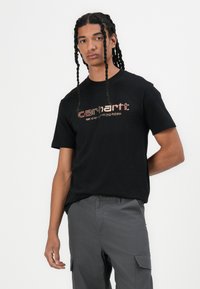 Black cotton t-shirt with "carhartt" logo in vibrant colors. Crew neck, short sleeves, worn with gray cargo pants. Plain design.