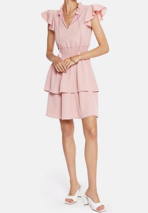 Frau trägt ein rosa Stufenkleid mit kurzen Ärmeln und Rüschendetails, einem gerafften Taillenbereich und weißen offenen Blockabsatz-Sandalen.