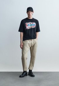 Νέος άνδρας που φοράει μαύρο γραφικό μπλουζάκι, μπεζ παντελόνι, μαύρες μπότες και μαύρο καπέλο, στέκεται μπροστά σε ένα απλό ανοιχτό γκρι φόντο.