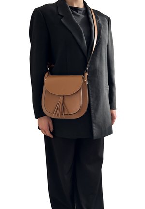 Persona che indossa un blazer nero e pantaloni con una borsa a tracolla in pelle marrone caratterizzata da un cappuccio e un dettaglio con nappina.