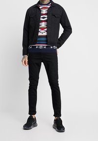 Czarna dżinsowa kurtka nałożona na sweter w wielokolorowy wzór, połączona z czarnymi jeansami slim-fit i czarnymi trampkami. Casualowy, warstwowy strój.