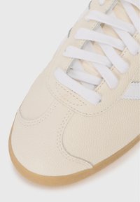 Crème-kleurige sneaker met een bovenlaag van textuurleer, witte veters en een gumrubberen zool. Beschikt over een eenvoudig ontwerp en subtiele stikseldetails.