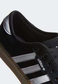 Černá semišová teniska s třemi bílými pruhy, černými tkaničkami a podrážkou z gumové pryže; horní část s texturou a polstrovaným límcem.