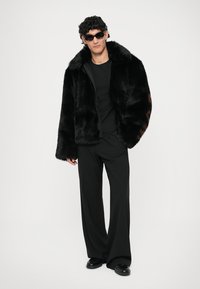 Ung mand iført sort faux fur-jakke, sort t-shirt, brede sorte bukser, loafers og sorte solbriller, stående mod hvid baggrund.