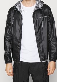 Svart lättviktsjacka med glänsande yta, justerbar huva och dragkedjefickor, med grå randig foder och logodetalj.