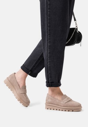 Mocassins en daim beige avec une semelle basse et épaisse et un détail de bande à l'avant, associés à un jean foncé retroussé et un sac à main noir.