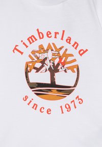 T-shirt blanc avec un motif graphique d'un arbre dans un emblème circulaire. Le texte "Timberland" et "depuis 1973" sont en orange.