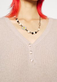 Beige pletený svetr s výstřihem do V a čtyřmi perleťovými knoflíky. Dekorativní náhrdelník obsahuje různé korálky a květinový akcent.