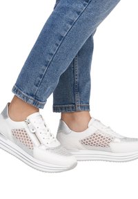 Weiße Sneakers mit Mesh-Panels, silbernen Akzenten und einem Reißverschluss. Die Sohle ist dick und geschichtet, kombiniert mit blauen, hochgekrempelten Jeans.