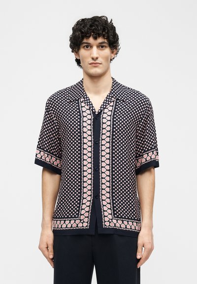 Νέος άνδρας με σγουρά μαλλιά, που φοράει κοντομάνικο μαύρο πουκάμισο με ροζ καρδιές, στέκεται μπροστά σε ένα απλό φόντο.