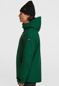 Groene winterjas met capuchon, voorzien van een zwarte borstzak en logo-detail. De stof lijkt glad, en de manchetten hebben een gestructureerde afwerking.