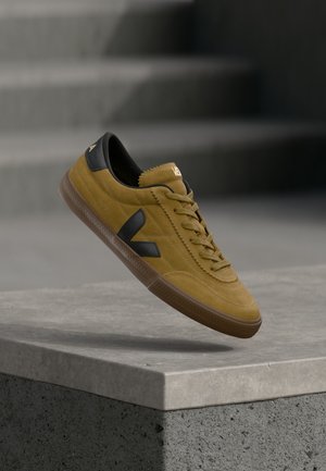 Żółty zamszowy sneaker z czarnymi skórzanymi akcentami, teksturowaną powierzchnią, płaskimi sznurowadłami i brązową gumową podeszwą, z logo na języku.
