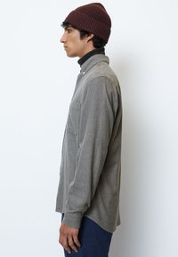 Button-up shirt in textured gray fabric, side view. Features a patch pocket, long sleeves, and subtle collar. Worn with a brown beanie.