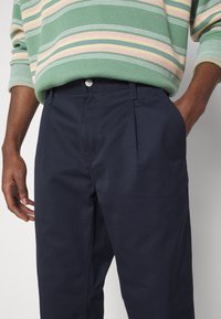 Granatowe spodnie z teksturowanego materiału, z przodu z zakładkami i zapięciem na guzik. Ręka w kieszeni, noszone z pastelowym swetrem w paski.