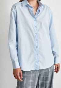 Femme portant une chemise bleu clair à boutons et un pantalon à carreaux gris, debout, les bras détendus le long du corps, devant un fond uni.