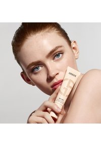 Beige tube med Armani Skin Tint holdt mot en models ansikt, med en matt finish og preget logo. Modellen har klar hud og lett sminke.