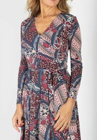 Kobieta w sukience z długim rękawem w bordowe, granatowe i kremowe kwiatowe oraz geometryczne wzory, mająca pomalowane na czerwono paznokcie i srebrny pierścionek.