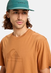 Ruskea puuvillapaita, jossa on hillitty Quiksilver-logo, yhdistettynä tealväriseen lippikseen, jossa on kaareva lippa ja ommeltuja yksityiskohtia.
