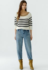 Pull rayé crème, noir et beige, avec un col large et des manches trois-quarts, associé à un jean taille haute bleu clair et des bottes beiges.