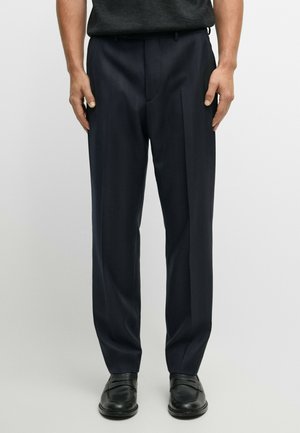 Man wearing dark navy dress pants and black leather loafers standing against a plain light background.