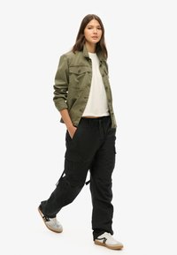 Militair-stijl olijfgroene jas, cropped witte t-shirt, zwarte cargo broek en grijze sneakers met zwarte strepen. Eenvoudig, functioneel ontwerp.