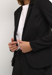 Schwarze Blazer mit taillierter Passform, ausgestattet mit einem einzelnen Knopf und glattem Stoff. Wird über einem weißen T-Shirt mit rundem Ausschnitt getragen.