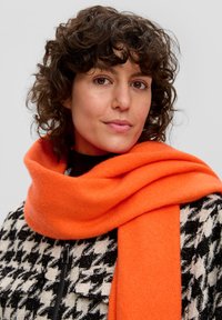 Écharpe en laine orange drapée autour du cou, associée à un blazer à motif pied-de-poule noir et blanc, doté d'une fermeture éclair.