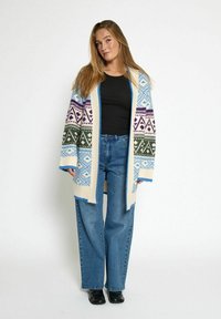 Cardigan a maglia con motivi geometrici nei colori blu, verde e viola, su una base crema e con bordi a coste. Abbinato a jeans a vita alta.